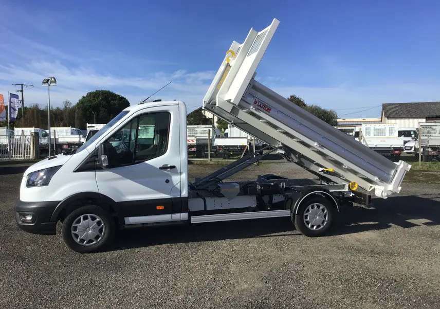 Vue de profil droit d’un Ford Transit blanc avec benne relevée et bras de levage visible sur fond extérieur.
