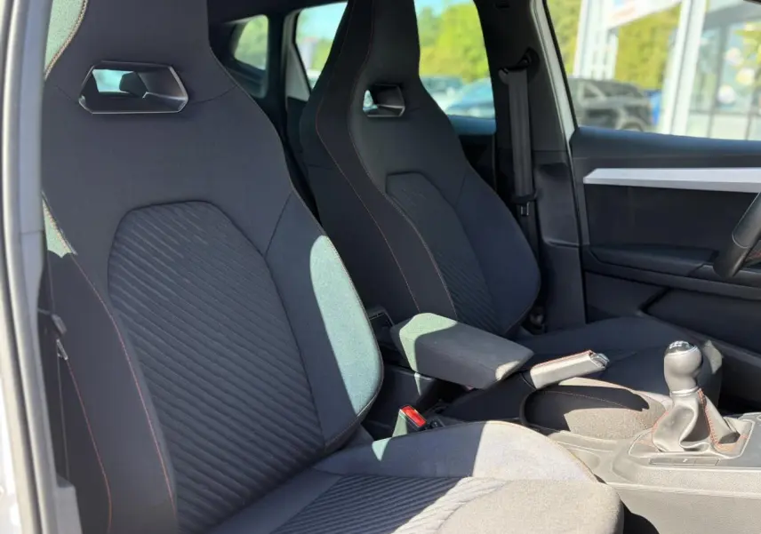 Vue intérieure côté conducteur de la SEAT Ibiza TSI 115 FR 2025, sièges sport noirs avec surpiqûres rouges et levier de vitesse manuel.