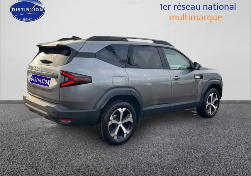 SUV Dacia Bigster gris schiste en 3/4 arrière droit avec jantes alliage diamantées et barres de toit visibles.