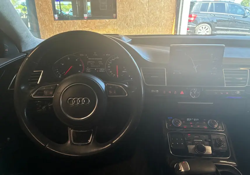 Vue intérieure centrée sur le volant et le tableau de bord d'une Audi A8 noire, avec écran de navigation et commandes visibles.