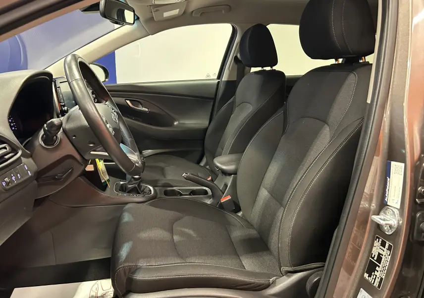 Vue intérieure côté conducteur de la Hyundai i30 SW T-GDI 120CH en Silky Bronze Metal, sièges tissu noirs avec volant cuir.