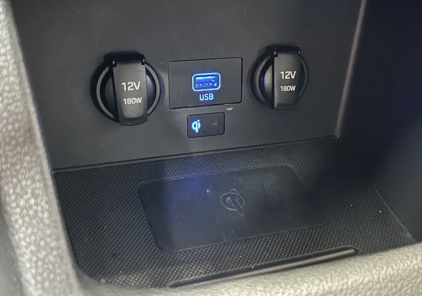 Gros plan sur la console centrale avec prises 12V, port USB bleu et zone de charge sans fil dans une Hyundai i30 SW bronze.