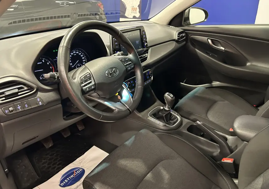 Vue intérieure de la Hyundai i30 SW 2022 côté conducteur, montrant le volant cuir, la boîte manuelle et les sièges tissu noirs.