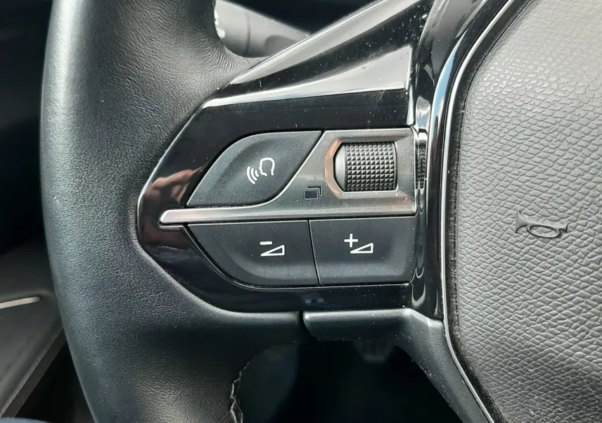 Gros plan sur les commandes gauche du volant en cuir noir du Peugeot 3008 gris Amazonite, boutons de volume et appel vocal.