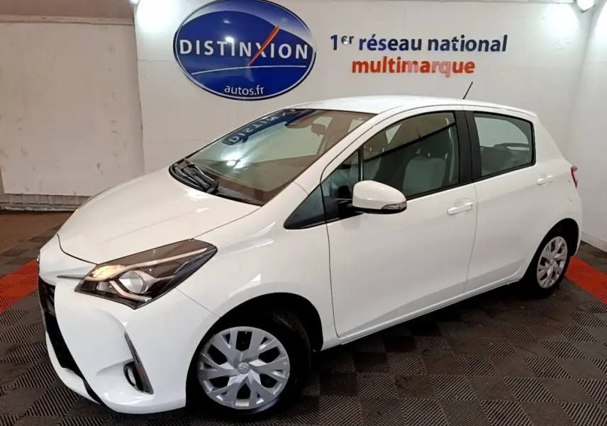 Toyota Yaris blanche vue 3/4 avant droit, avec phares allumés et jantes en alliage visibles.