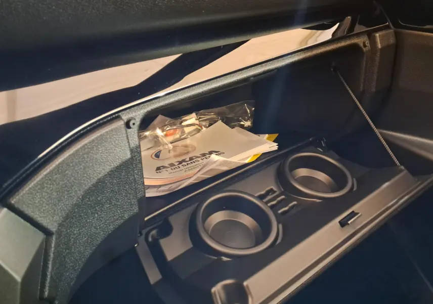 Gros plan sur la boîte à gants ouverte d'une Aixam Ambition rouge, avec porte-gobelets et documents visibles à l'intérieur.