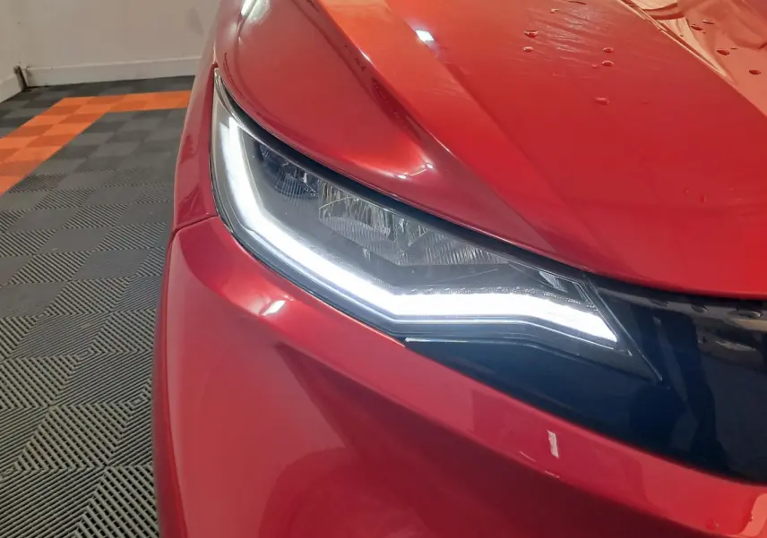 Gros plan sur le phare avant gauche allumé d'une Aixam Ambition rouge 2025 dans un garage au sol carrelé.