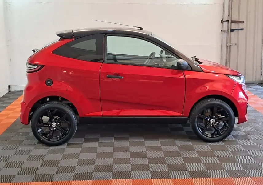 Profil côté gauche de l'AIXAM AMBITION rouge 2025 avec toit noir et jantes noires sportives dans un intérieur lumineux.