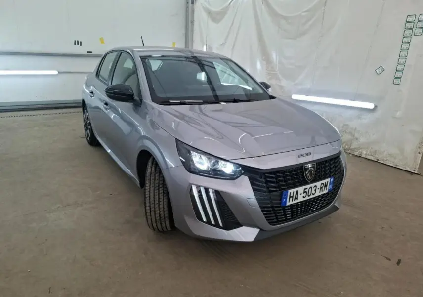 Peugeot 208 gris clair vue 3/4 avant droit, phares allumés et calandre noire distinctive, dans un garage.