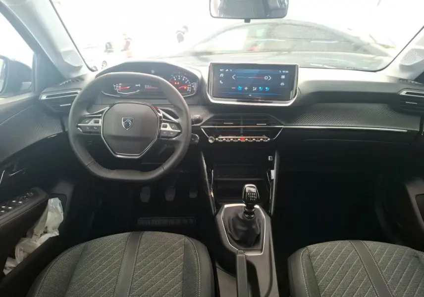 Vue intérieure frontale du tableau de bord et volant de la Peugeot 208 2024, avec boîte manuelle et sièges gris.