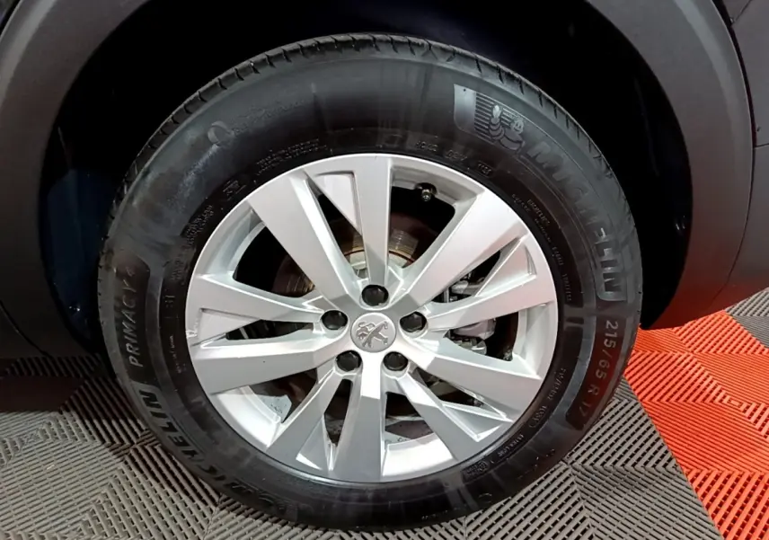 Gros plan sur la roue avant droite d’un Peugeot 5008 noir, jante alliage argentée avec logo lion au centre.