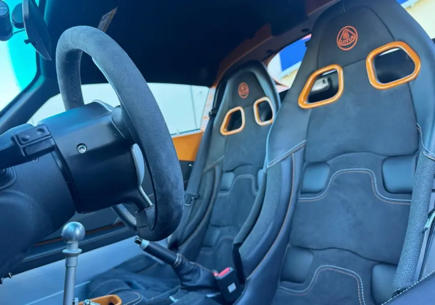Vue intérieure côté conducteur de la Lotus Elise 3 Cup 250 2021, sièges sport noirs avec surpiqûres orange et volant cuir.