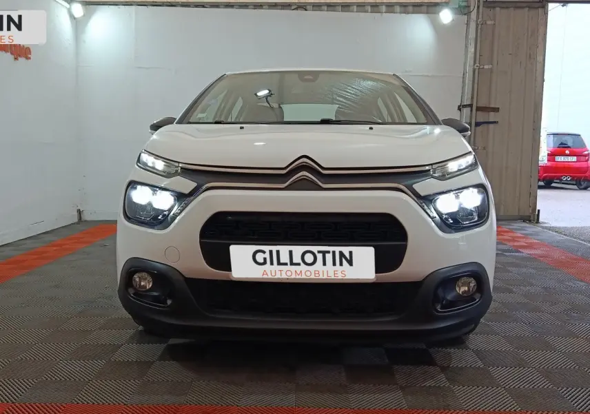 Vue de face d'une Citroën C3 blanche avec phares allumés dans un garage au sol carrelé rouge et gris.