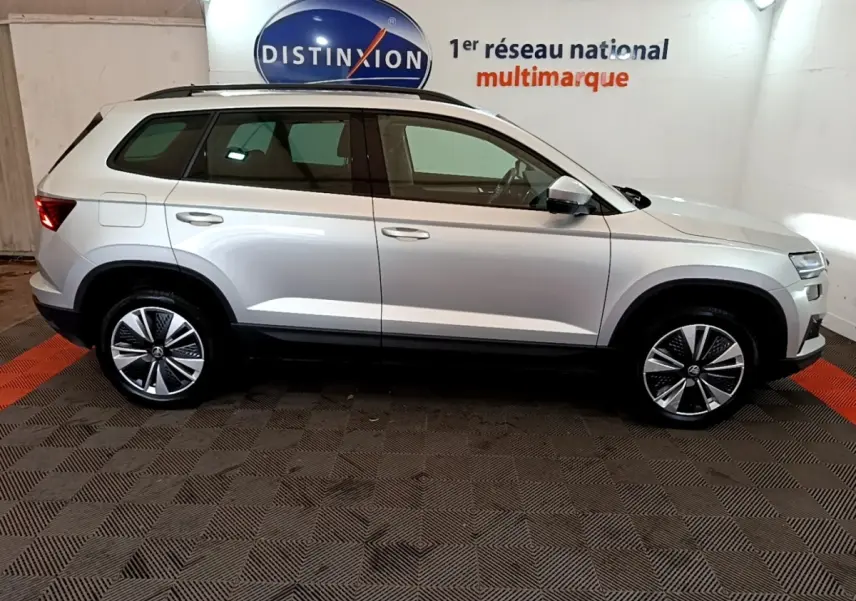 Vue latérale droite d’un Skoda Karoq gris clair 2022 avec jantes alliage et barres de toit noires en showroom.