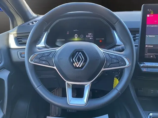 Volant noir de Renault Captur 2024 vu de face, avec commandes intégrées et tableau de bord numérique en arrière-plan.
