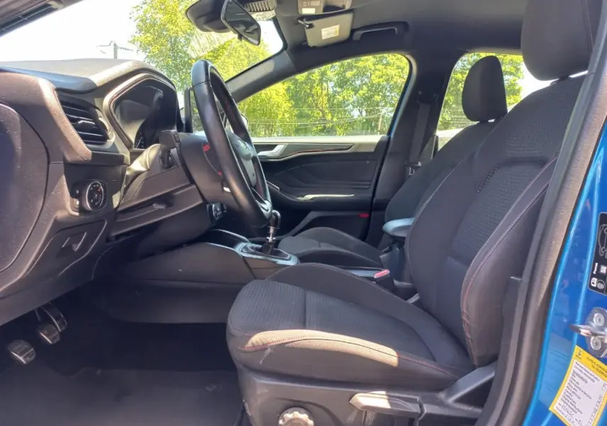Vue intérieure côté conducteur d'une Ford Focus bleu 2019, montrant les sièges noirs avec surpiqûres rouges et la boîte manuelle.