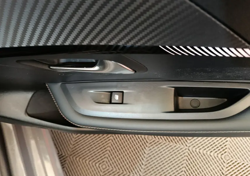 Gros plan sur la commande de lève-vitre côté conducteur de la Peugeot 208 gris foncé, avec insert décoratif en fibre de carbone.