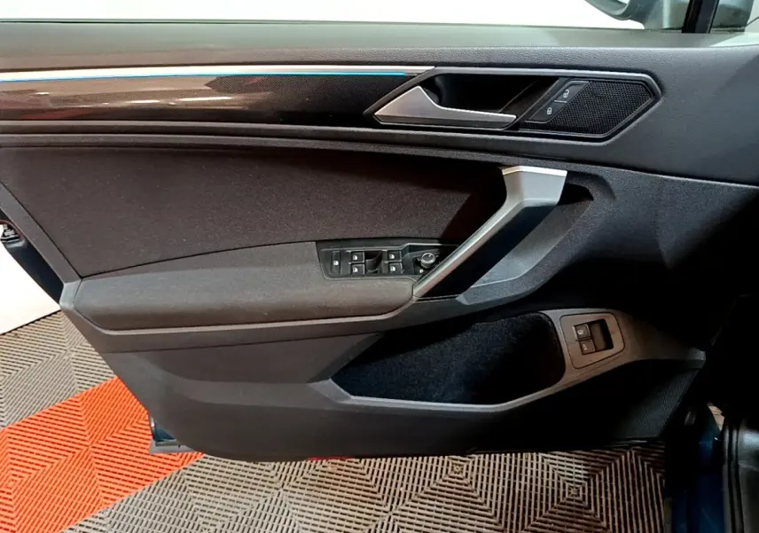 Vue intérieure côté gauche de la porte avant d’un Volkswagen Tiguan bleu, avec commandes électriques et garniture noire.