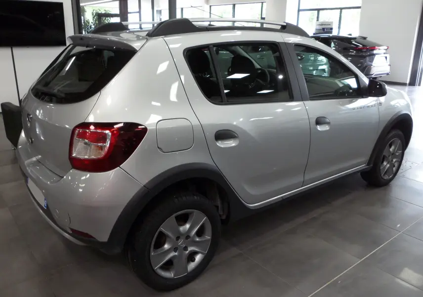 Vue 3/4 arrière droite d'une Dacia Sandero Stepway gris platine en intérieur, avec jantes alliage et barres de toit noires.