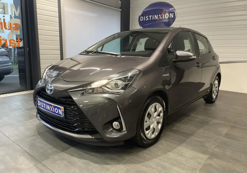 Toyota Yaris hybride gris foncé vue 3/4 avant droit en intérieur avec calandre noir laqué et phares allumés.