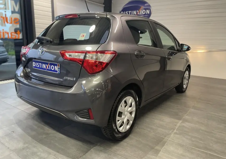 Toyota Yaris hybride gris foncé vue 3/4 arrière droit en intérieur avec jantes à enjoliveurs blancs.