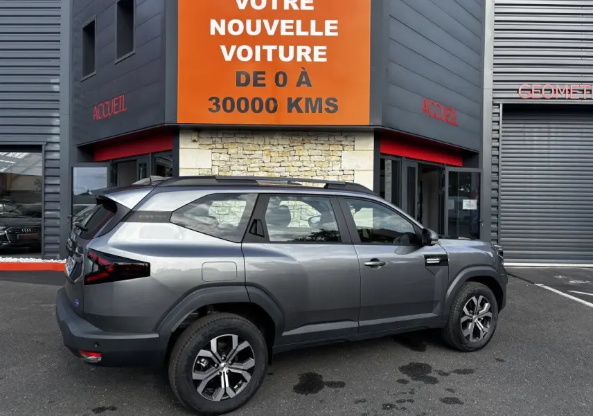 Vue de profil côté gauche d'un Dacia Bigster gris foncé avec rails de toit noirs garé devant un bâtiment commercial.