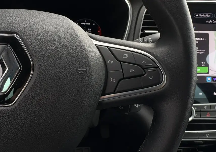 Gros plan sur le volant noir de la Renault Mégane Break 2024 avec commandes intégrées et écran tactile GPS en arrière-plan.