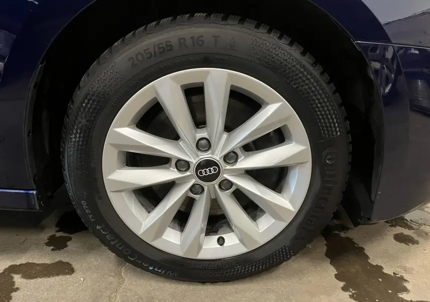 Gros plan sur la roue avant droite avec jante argentée et logo Audi sur une carrosserie bleu métallisé.