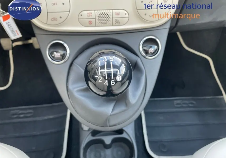 Le levier de vitesses manuel noir au centre de l'habitacle d'une Fiat 500 blanche, avec la console et les commandes climatiques visibles.
