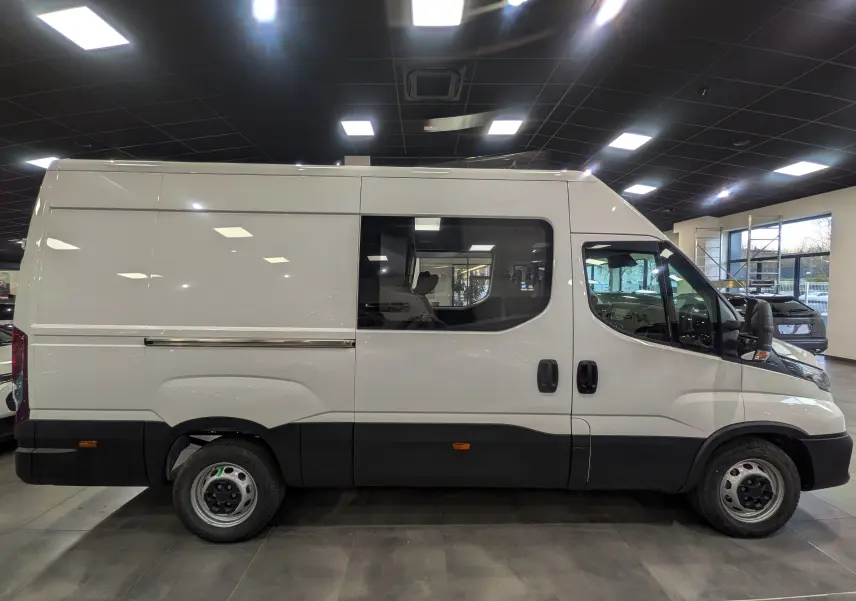 Vue latérale droite d'un fourgon Iveco Daily blanc 2025 avec fenêtre centrale et bas de caisse noir dans un showroom.