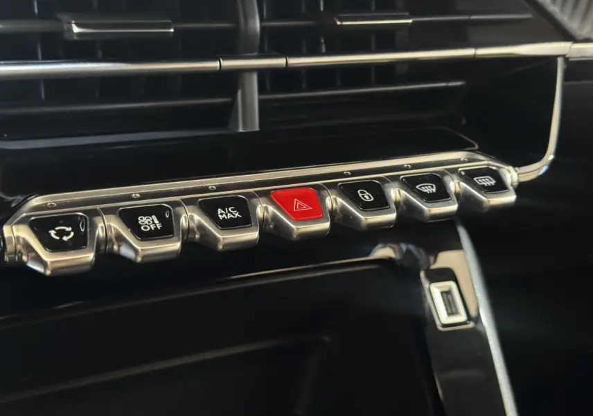 Gros plan sur la rangée de boutons métalliques du tableau de bord de la Peugeot 208 jaune, avec bouton rouge d’alerte au centre.