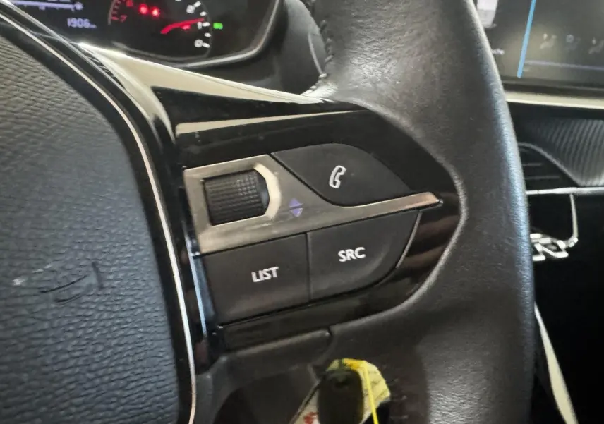 Gros plan sur les commandes gauche du volant cuir noir d’une Peugeot 208 2023, avec tableau de bord en arrière-plan.