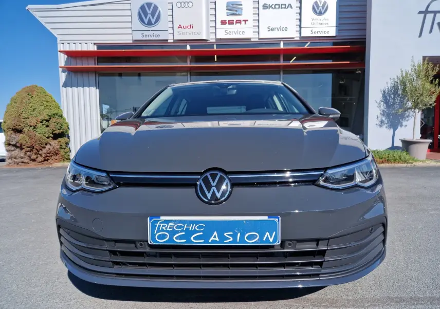 Vue frontale d'une Volkswagen Golf A8 gris Urano 2023 avec plaque d'occasion devant un garage multimarques.