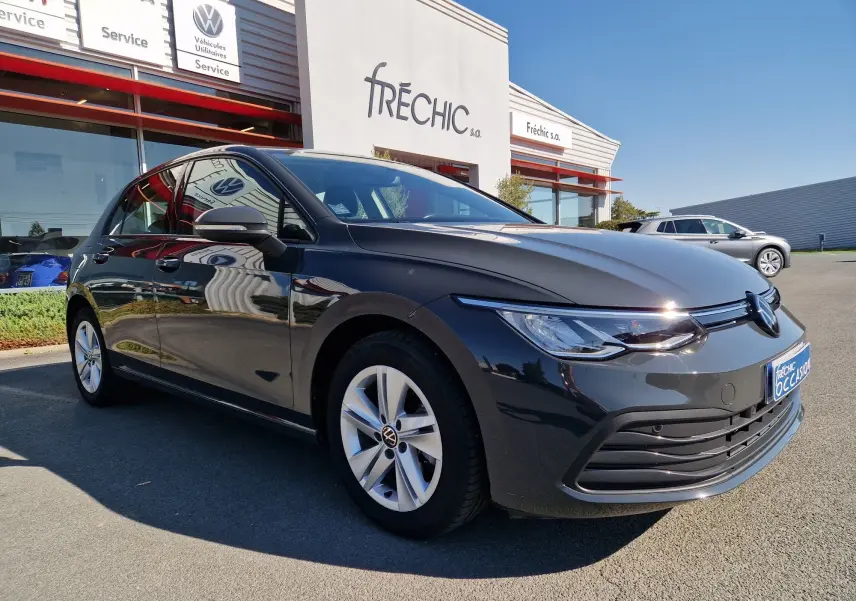 Volkswagen Golf A8 gris Urano vue 3/4 avant droit, avec jantes alliage et calandre moderne, stationnée devant un concessionnaire.