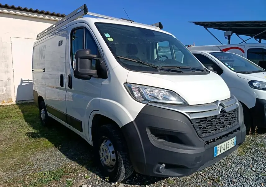 Vue 3/4 avant d'un utilitaire blanc Citroën Jumper L1H1 avec galerie de toit et pare-chocs noir sur un sol gravillonné.