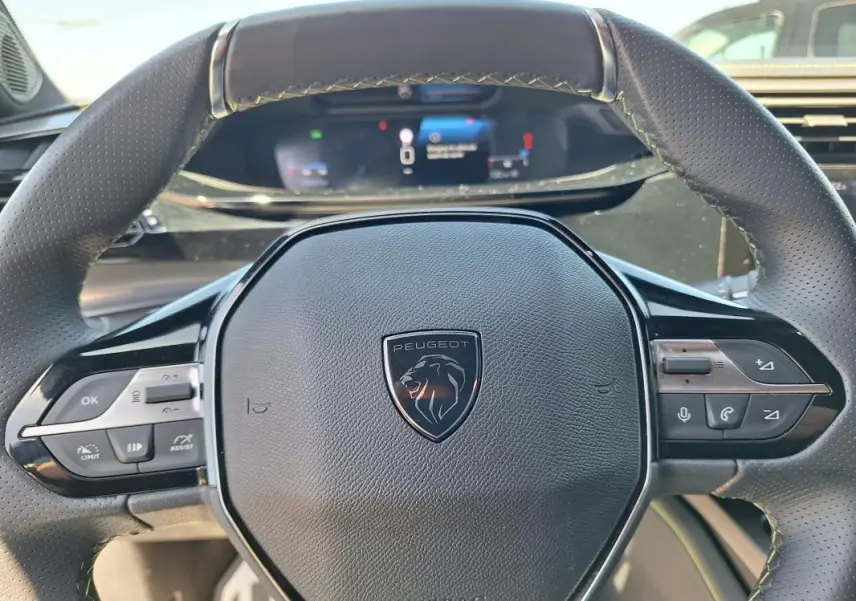Vue rapprochée du volant cuir noir de la Peugeot 308 Hybrid 145 e-DCS6 GT avec commandes intégrées et tableau de bord numérique flou.