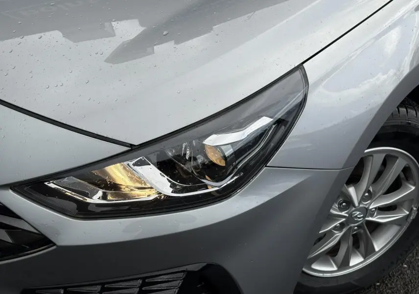 Gros plan sur le phare avant droit allumé et la jante alliage d’une Hyundai i30 SW T-GDI 120 Modern gris argent sous la pluie.