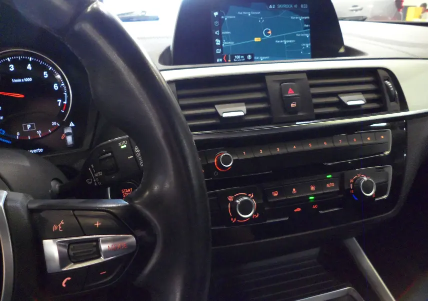 Vue intérieure centrée sur le tableau de bord et le volant noir d'une BMW Série 1 F21 LCI avec écran de navigation allumé.