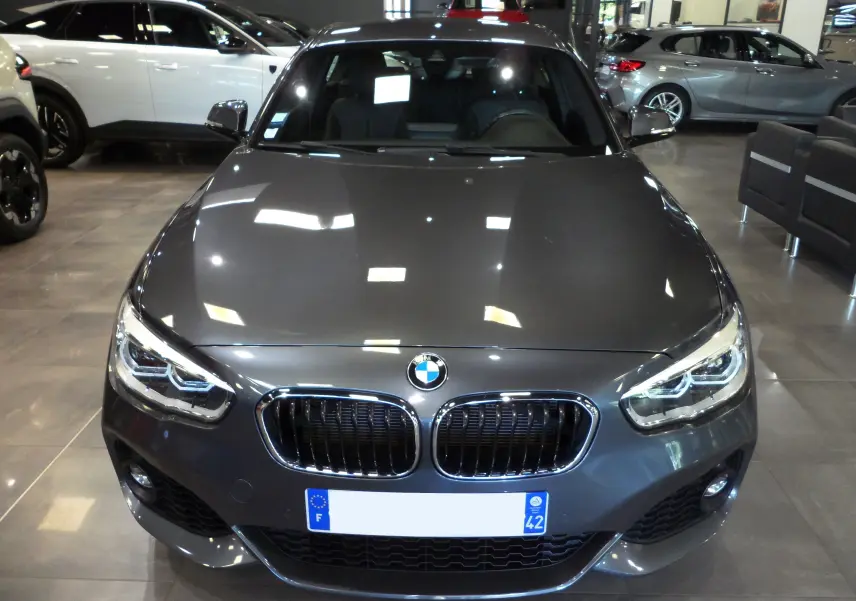 Vue frontale d'une BMW Série 1 F21 LCI gris métallisé en showroom, mettant en valeur sa calandre double et ses phares LED.