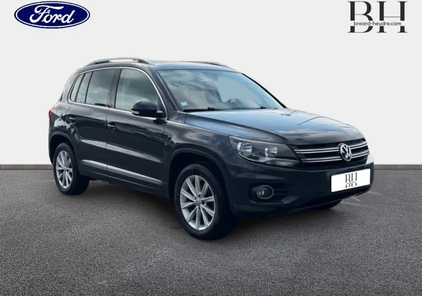 Volkswagen Tiguan gris poivre en 3/4 avant droit, avec jantes alu et barres de toit visibles.