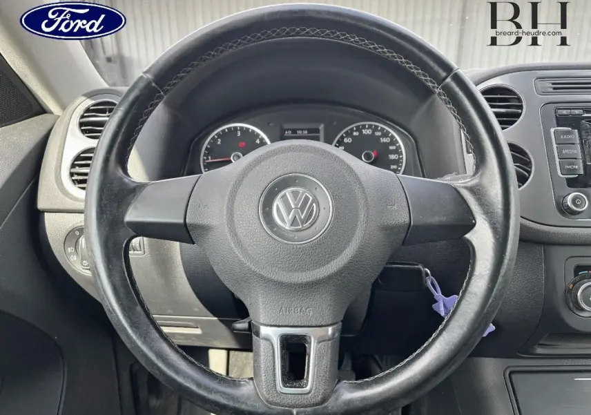 Vue rapprochée du volant noir avec logo Volkswagen au centre, tableau de bord et commandes visibles en arrière-plan.