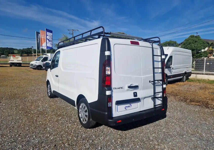 Vue 3/4 arrière droite d’un Renault Trafic blanc avec échelle et galerie noire sur toit, sur un terrain extérieur.