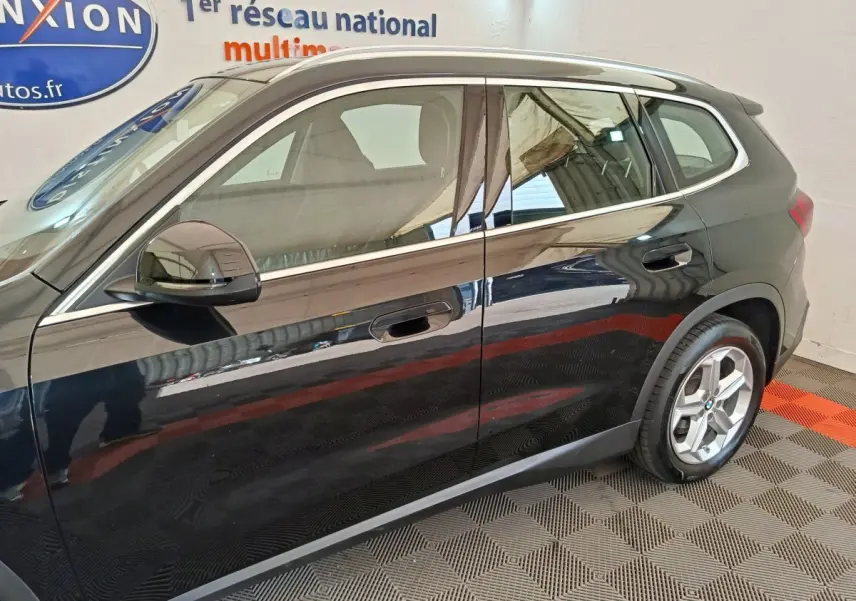 Vue latérale droite d’un BMW X1 noir 2023 en intérieur, avec rétroviseur rabattable et jantes visibles.