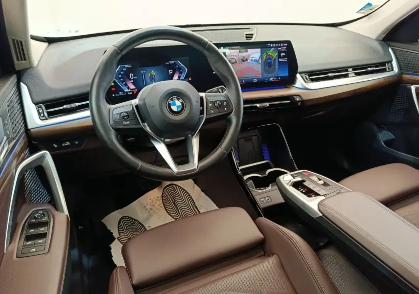Vue intérieure avant du BMW X1 2023 avec volant cuir, écran tactile large et finitions bois marron.