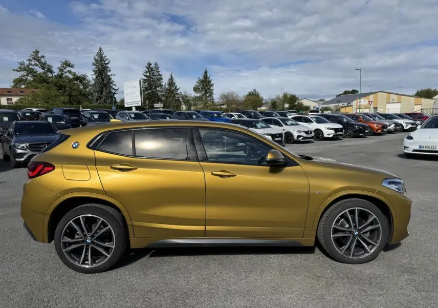 Profil côté gauche d'un BMW X2 jaune métallisé avec jantes M sport noires sur un parking extérieur.