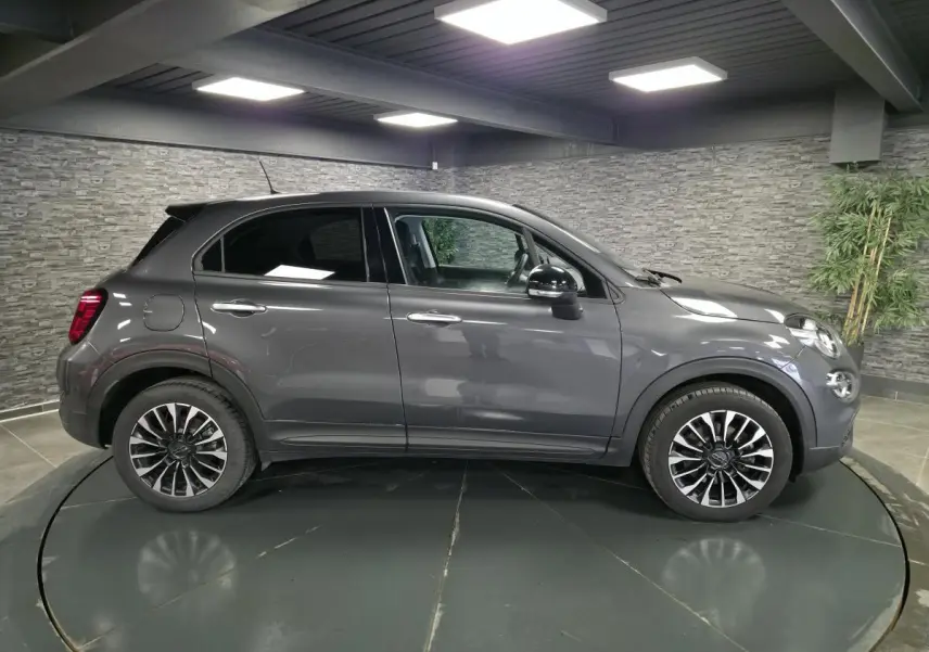 Profil droit d'un Fiat 500X gris avec jantes alliage 17'' et vitres surteintées en intérieur showroom.