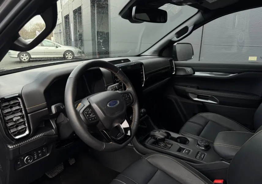 Vue intérieure côté conducteur du Ford Ranger 2025, tableau de bord noir avec surpiqûres orange et volant cuir multifonctions.