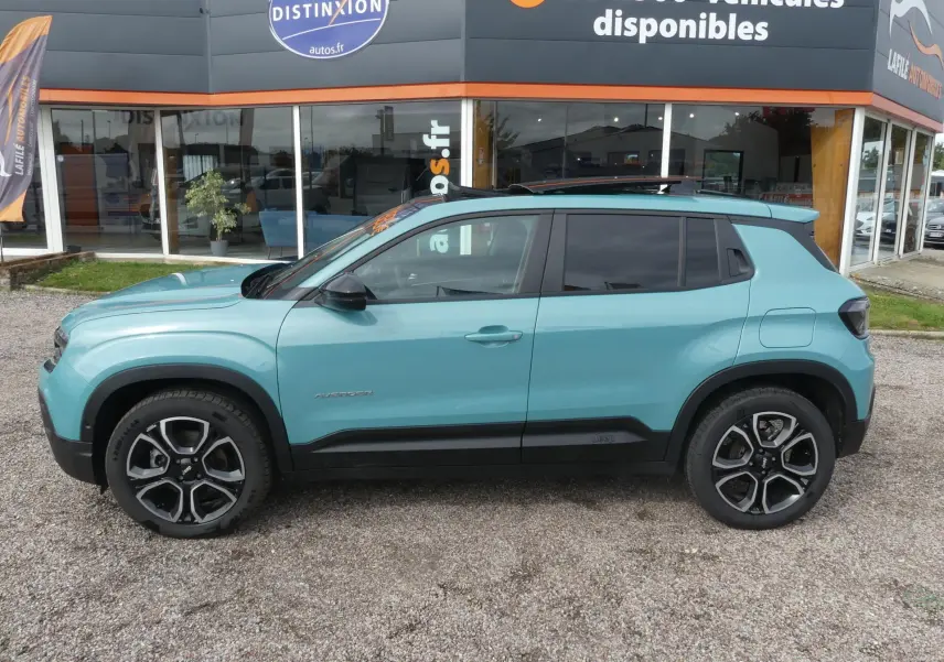 Profil côté gauche du Jeep Avenger bleu métallisé 2024 avec toit ouvrant et vitres surteintées stationné devant un showroom.