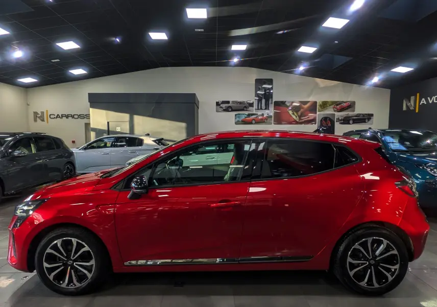 Profil côté gauche de la Renault Clio V rouge flamme 2025 en intérieur, avec jantes alliage et rétroviseurs noirs.