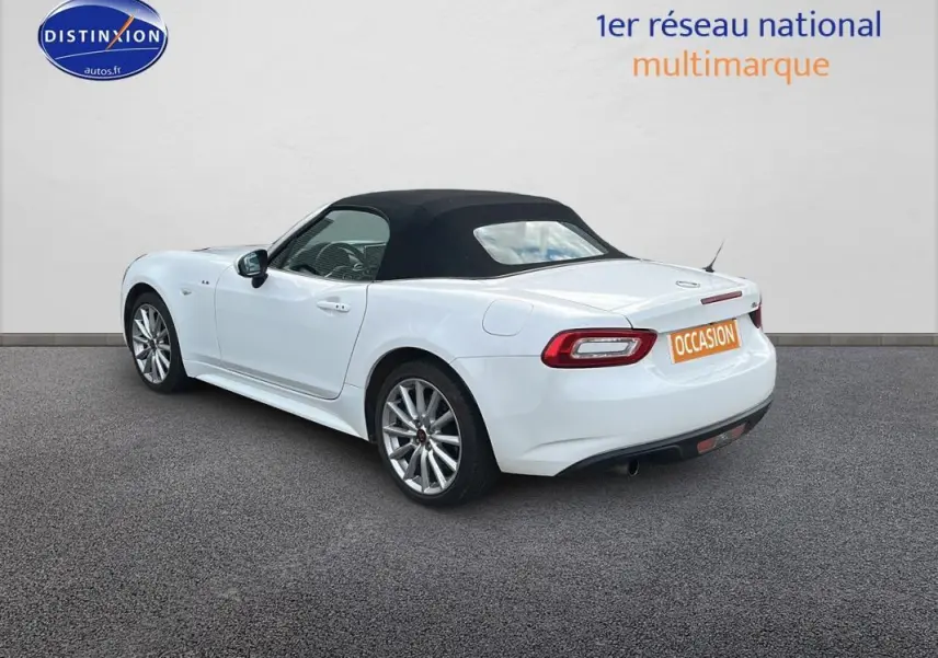 Fiat 124 Spider blanc vue 3/4 arrière côté gauche, capote noire fermée et jantes alliage 17 pouces visibles.